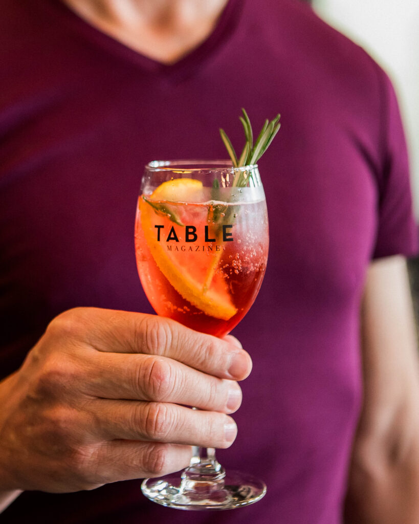 A man in a purple shirt holds a pink drink in a TABLE Magazine cocktail glass with a spring of rosemary sticking out of the top.