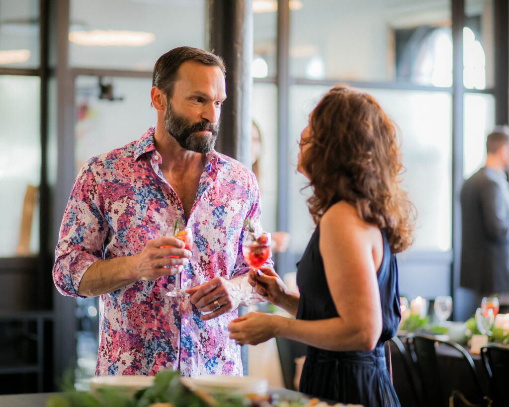 A man in a bright patterned shirt holds a red cocktail and talks to a woman in a blue dress also holding a red drink.