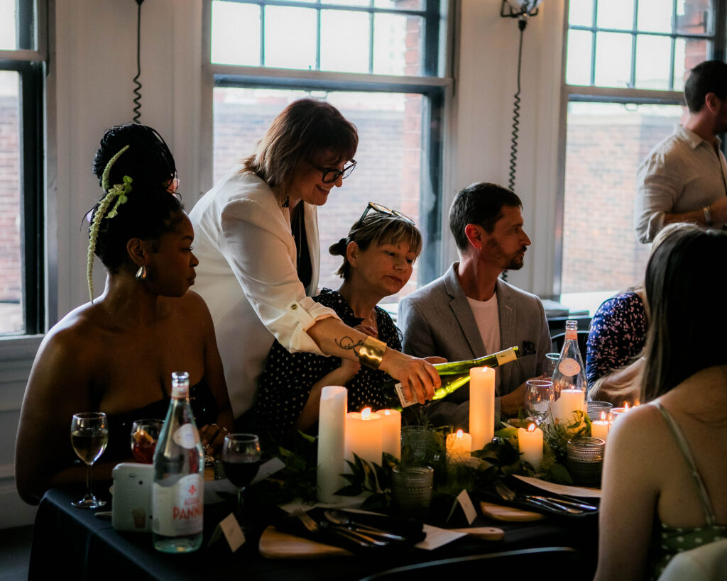 A woman in white pours wine for another woman seated at a long table with candles.