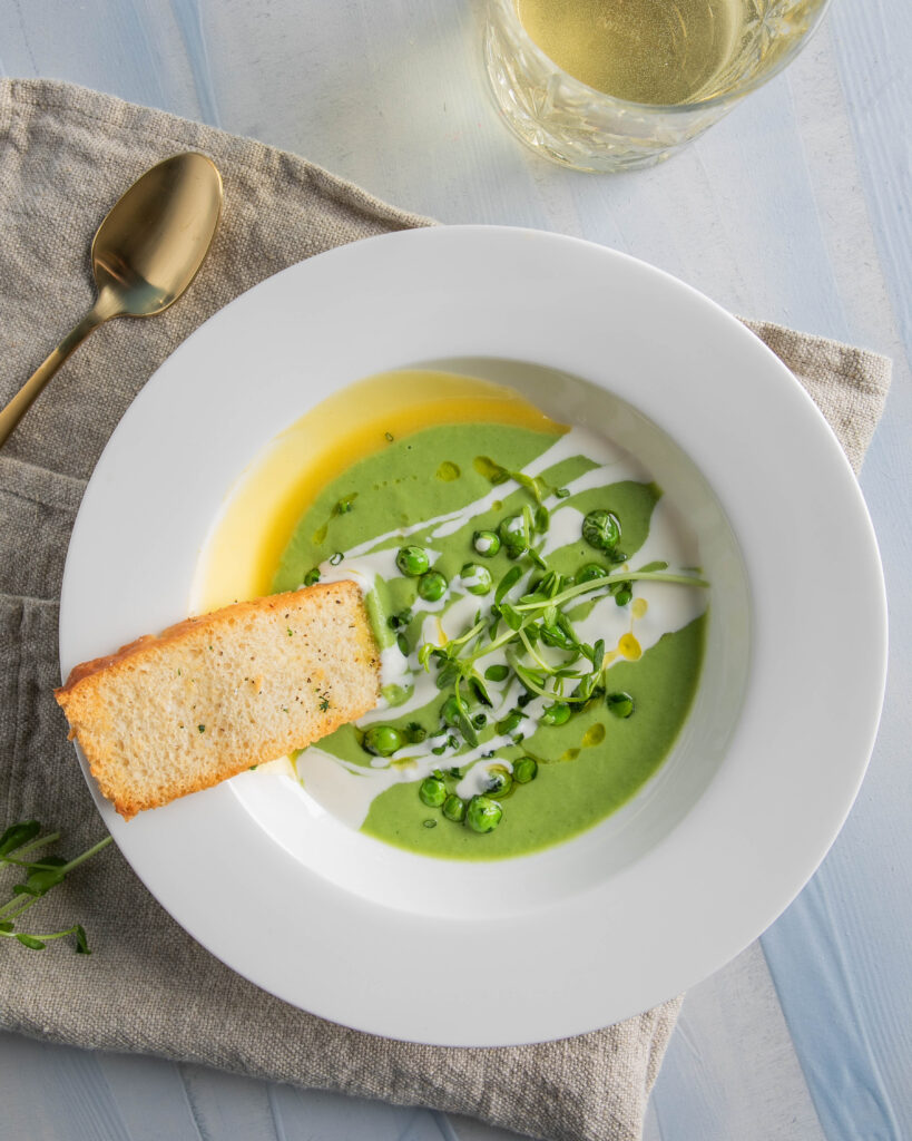 A white bowl holds a creamy green soup with a slice of bread on the brim of the bowl.