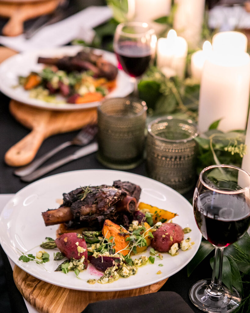 A white plate holds lamb and various vegetables on a table surrounded by wine glasses, greenery, and candles.