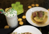 2024 Saint Patrick’s Day Menu Specials in Pittsburgh A plate of bangers and mash sits on a table along with a festive saint patrick's day creamy drink to the left and gold coins scattered throughout.