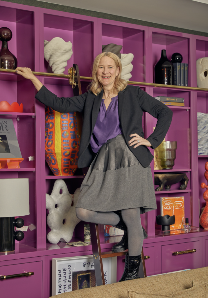Brenda Friday stands on a step ladder beside pink cabinetry in a black blazer, purple shirt, and grey skirt.