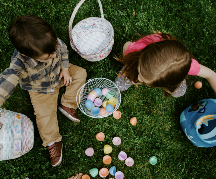 Two children sit on the grass and sort through their baskets after an Easter egg hunt with empty eggs sitting in front of them.