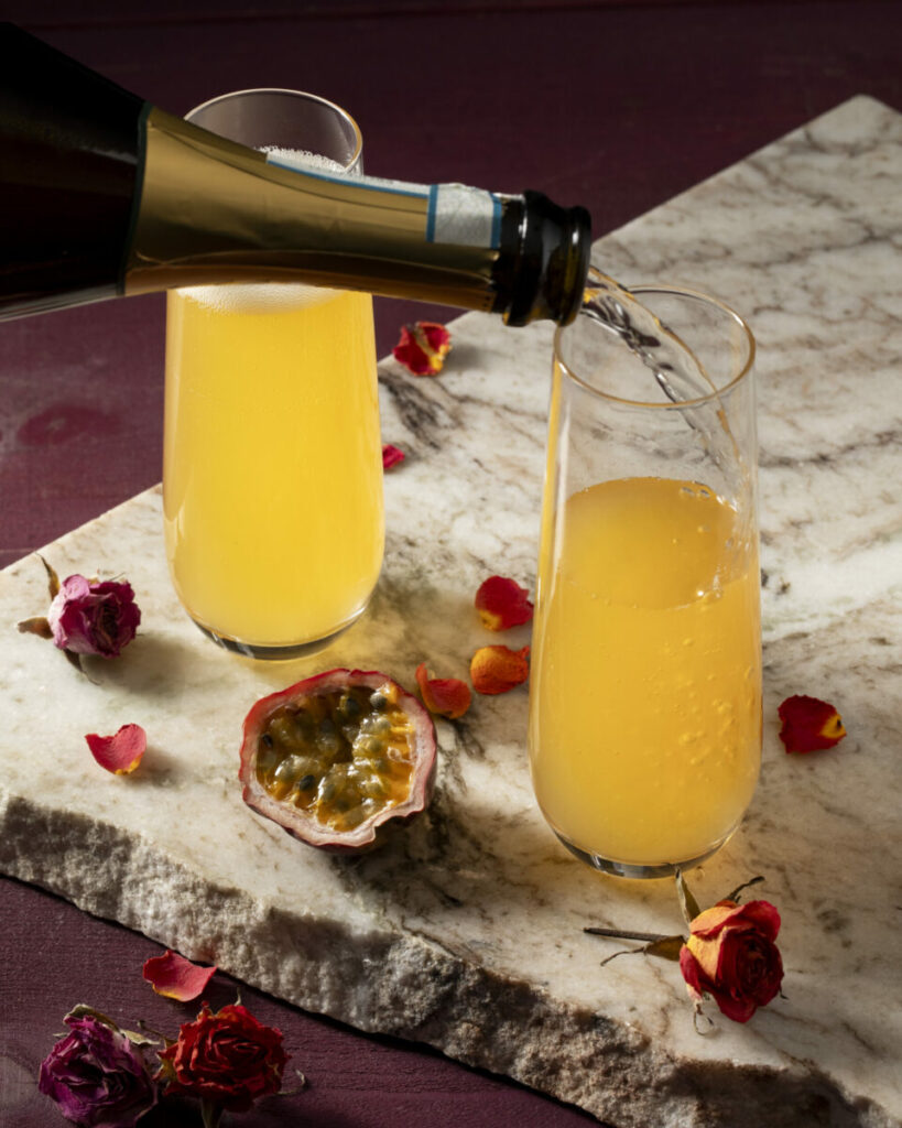 Two champagne flutes hold a yellow-orange colored drink as sparkling wine is poured into the glass on the right to make the More Adventurous Valentine's Day Cocktail.