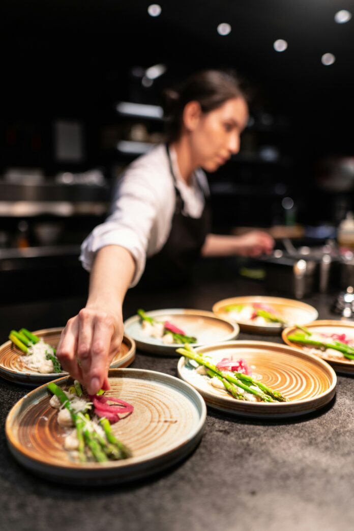 A woman chef in the kitchen in Pittsburgh garnishes plates with vegetables while her hand is in focus and her body is not.