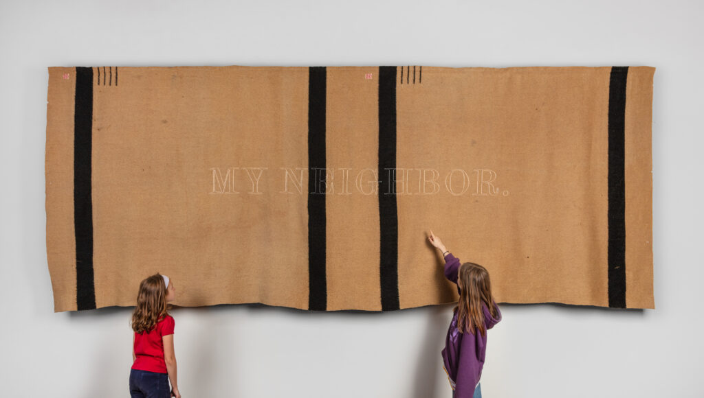 A brown and black quilt sits on a wall as two young girls point up at the words printed on it in white.