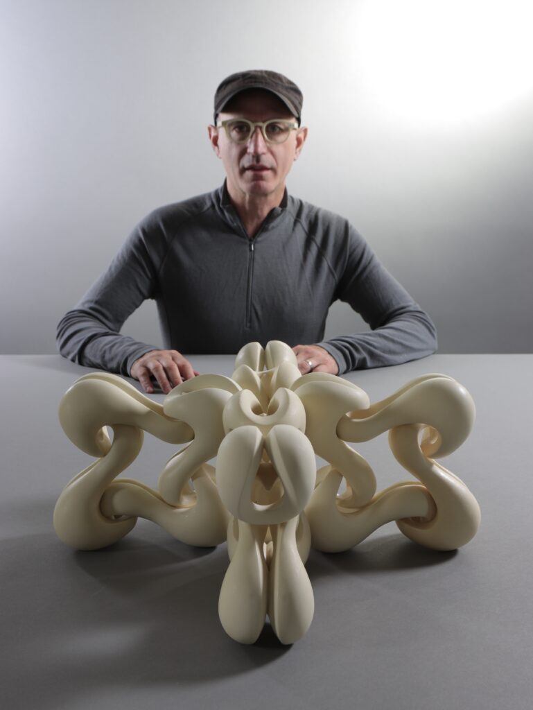 Sculptor Isaac Bower sits at a grey table with one of his art pieces sitting in front of him in the form of a spiral sculpture.