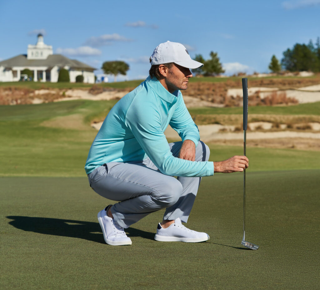 A man playing golf kneels down in his men's fashion clothing with a baseball cap on his head and golf club in hand.