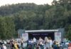 Allegheny County Summer Concert Series ’24 A rear view of people in lawn chairs on the grass enjoying a concert on the stage in the park.