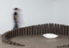 Contemporary Craft Explores Grief with New Exhibit “Hereafter” A variety of different sized candles sit in an oval shape on the floor around a pillow while a person walks in a blurred motion behind the candles.