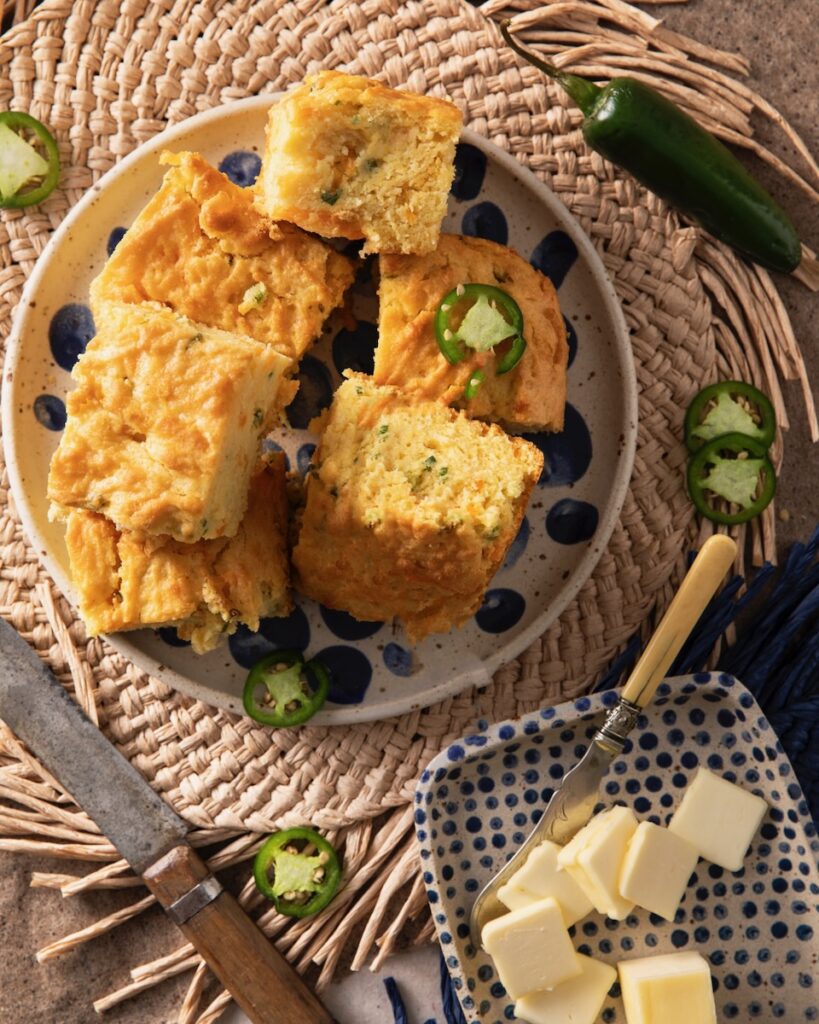 A plate of stacked cheddar cornbread sits beside a plate of butter slices and knives with jalapeños scattered throughout.