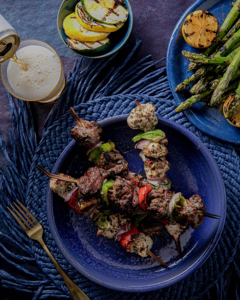 Four kabob sticks sit on a blue plate against a blue table cloth with a plate of asparagus, beer, and fork nearby.