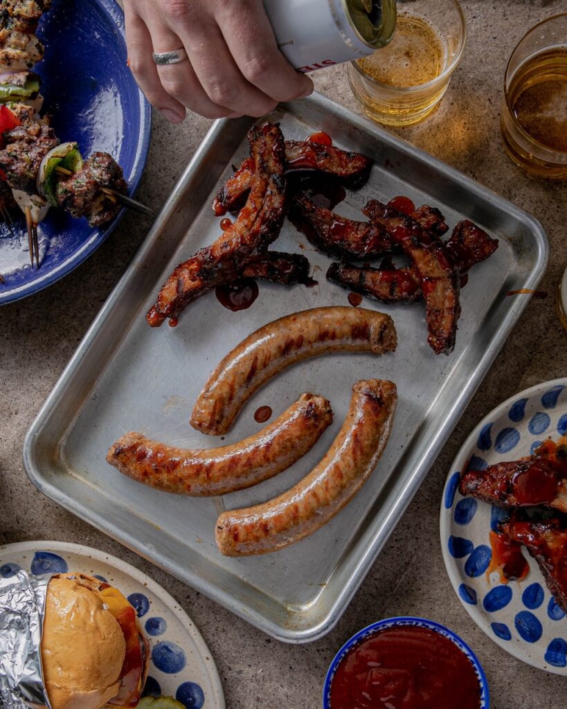 Various grilled sausages sit on a silver sheet pan with small plates and a beer glass sitting around the sheet pan. 