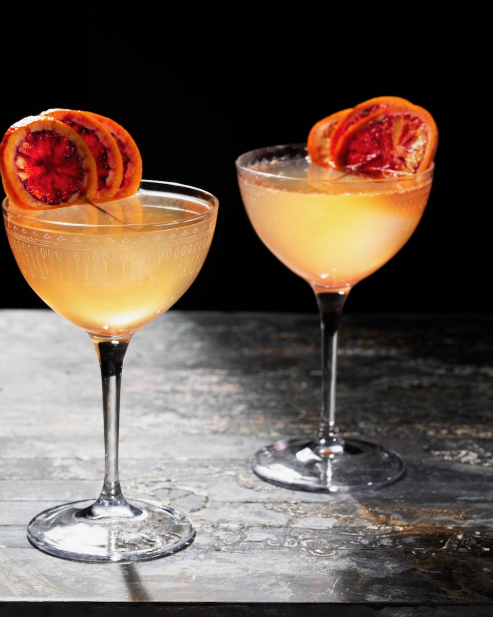 A yellow cocktail in a coupé glass with an orange slice in it on a black background 