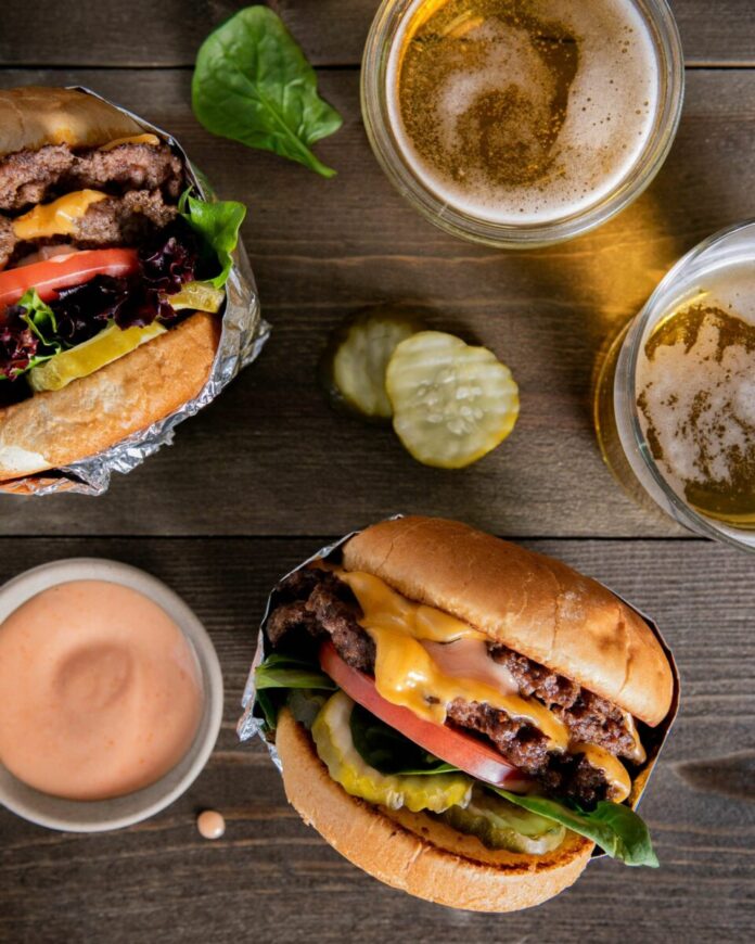 two hamburgers pickles an orange sauce and 2 cups of bear on a brown wooden table