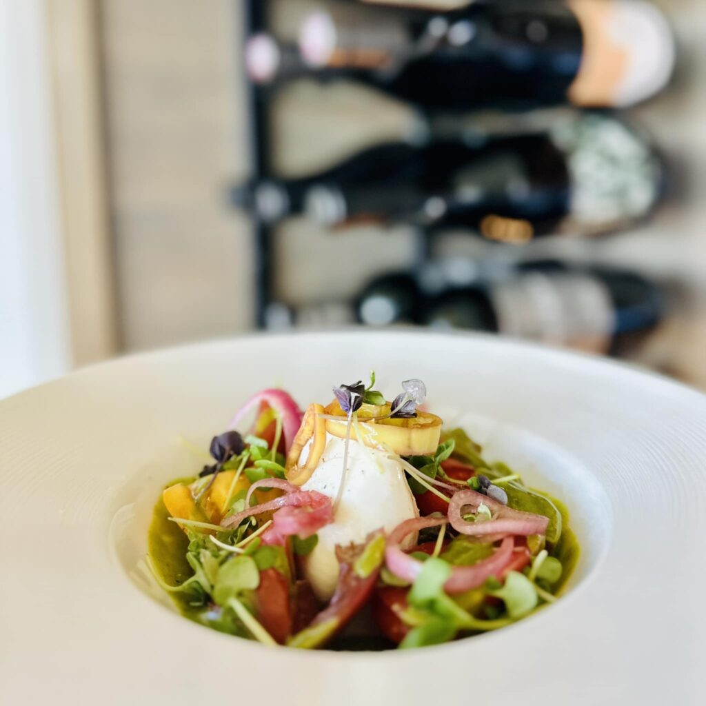 A small salad sits in a white round bowl in front of a wine rack.