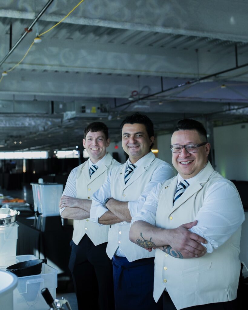 Three men in white vests and ties cross their arms in a line and smile at the camera. 