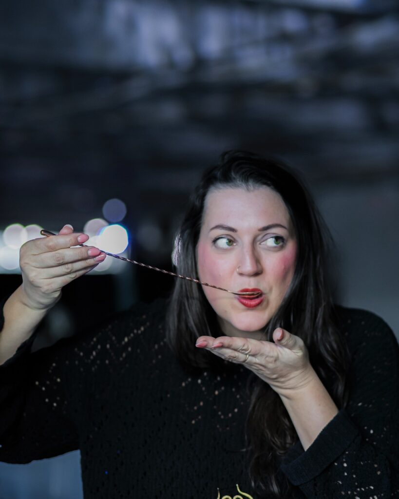 A woman with dark hair and lipstick tastes a cocktail with a long stirring spoon. 