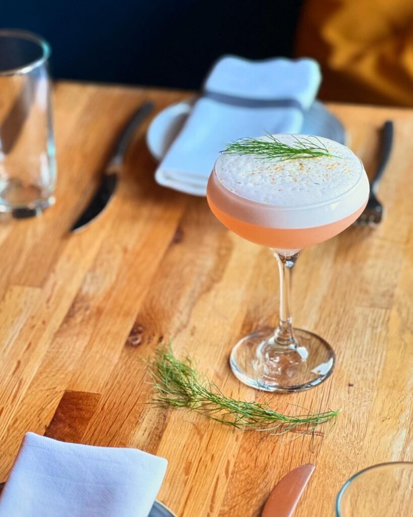 A cocktail with foam on top from Fig and Ash and topped with a sprig of green sits on a wooden table.