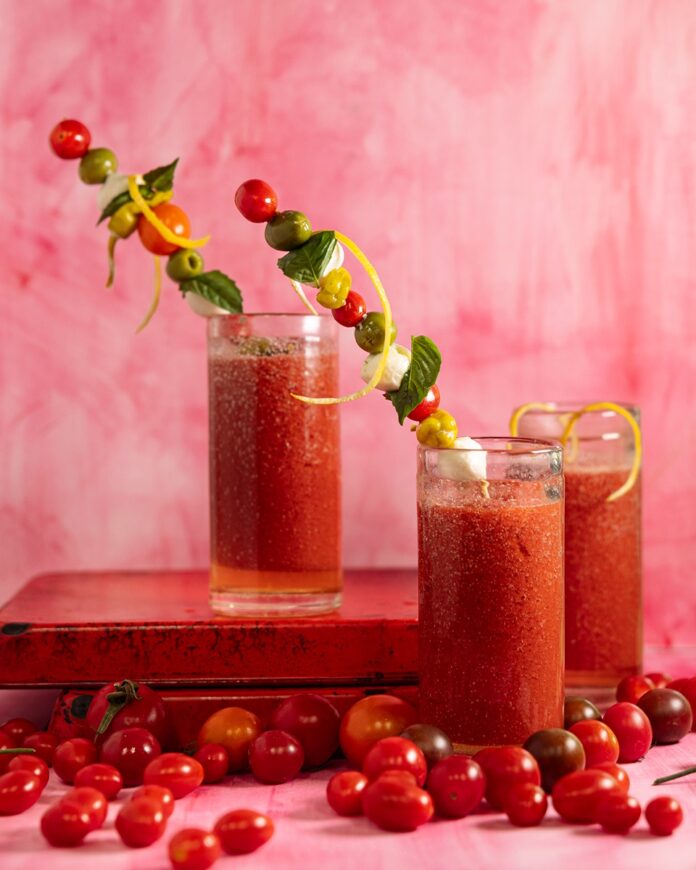 Garden-Fresh Caprese Bloody Mary Three tall glasses filled with a vibrant red tomato-based drink, likely Bloody Marys, each garnished with skewers featuring colorful cherry tomatoes, mozzarella balls, green olives, basil leaves, and lemon zest spirals.