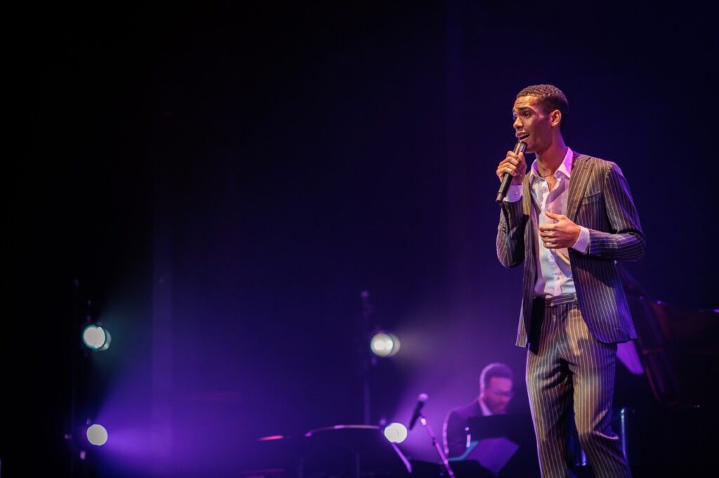 A man sings on a concert stage with a microphone in hand and purple lights throughout.