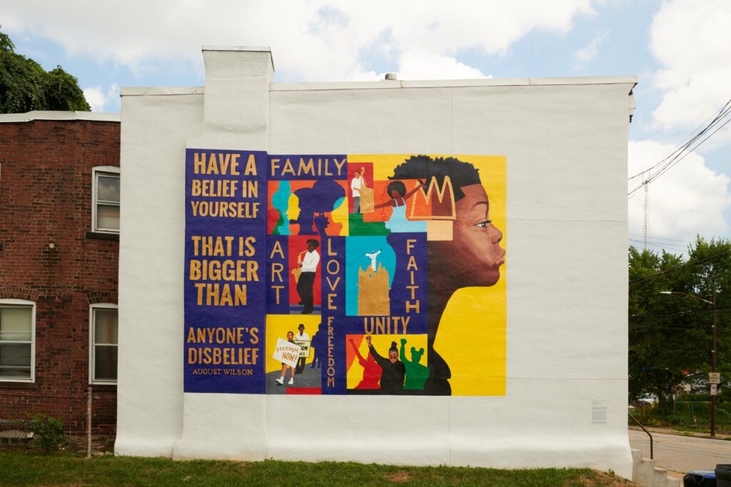 A white outside building in Pittsburgh is designed in an artwork in bold yellow and purple colors with a young Black child's face to the right of different phrases.