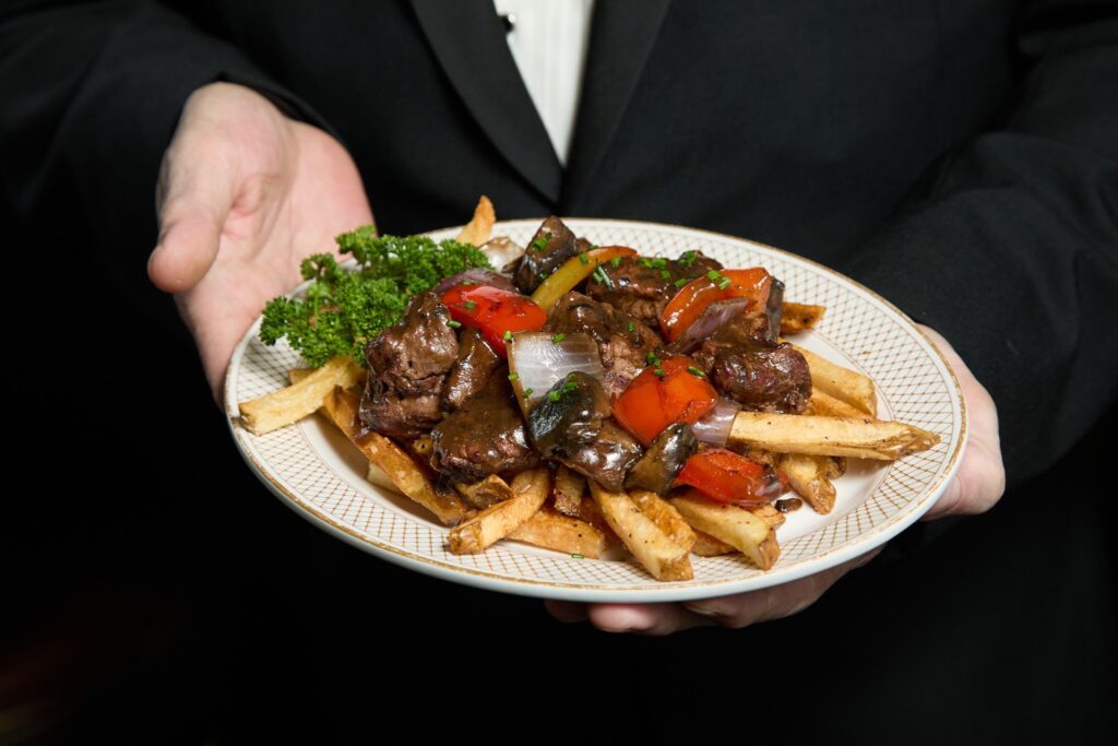 A man in a tuxedo holds out a white plate topped with fries and beef tips.