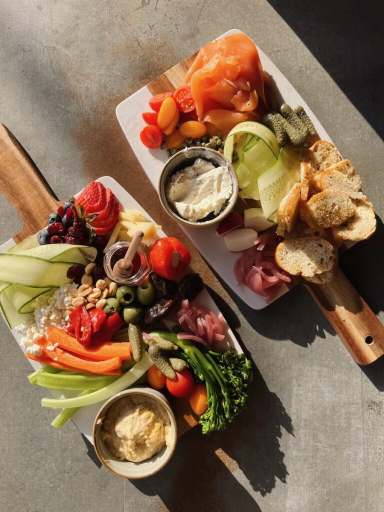 Two full charcuterie boards with vegetables, dips, cheese, and salmon sit on a table.