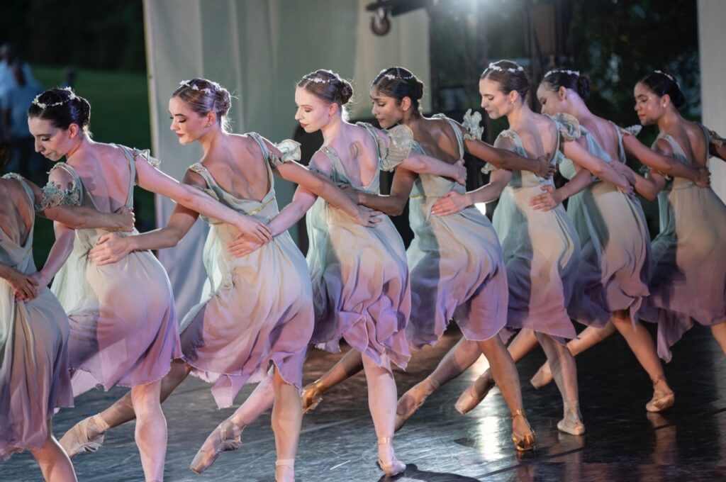 A line of dancers in purple and white dresses cross their arms behind each others back in Pittsburgh Ballet Theatre's under the stars production.