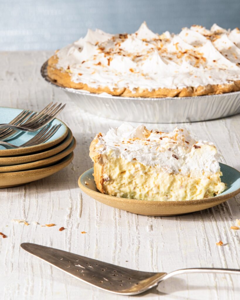 A slice of coconut cream pie from a sweet treat place in Pittsburgh sits in front of the whole pie, with a stack of plates and forks to the left and a spatula in front of the plate.