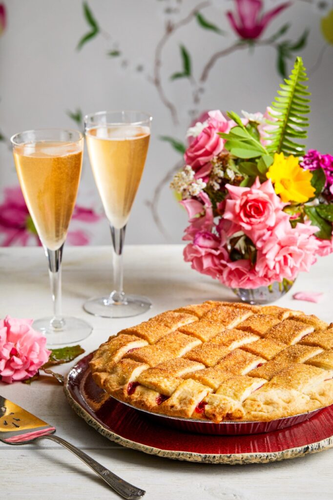 A lattice baked pie from a sweet treat eatery in Pittsburgh. Behind the pie sits two champagne glasses and a flower display in pink.