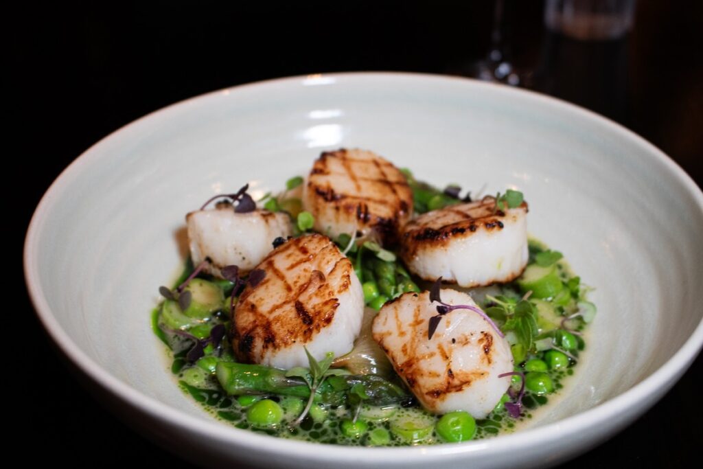 A white bowl holds a green pea base topped with grilled scallops.