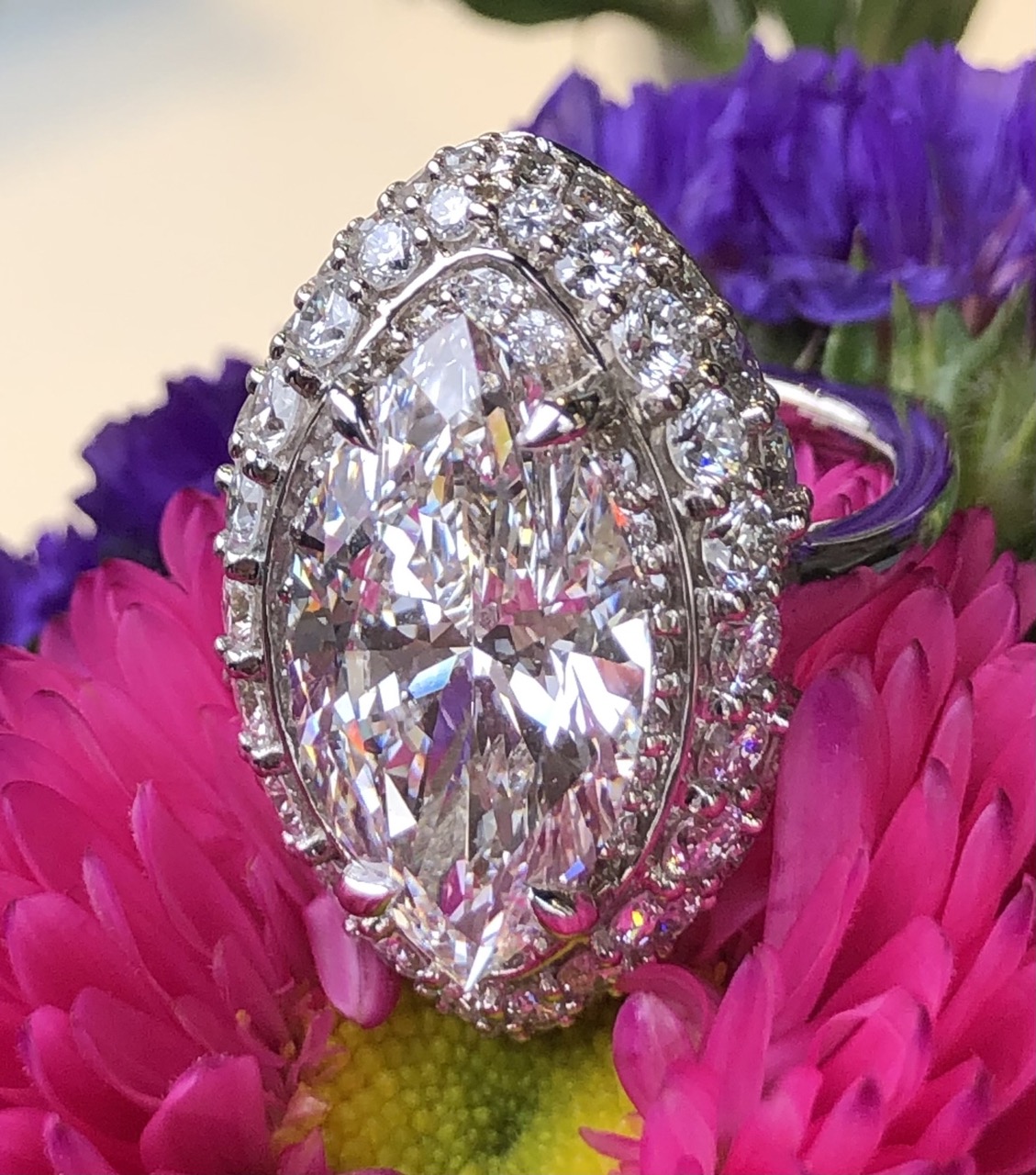 A custom ring in an oval diamond shape sits against pink and purple flowers.