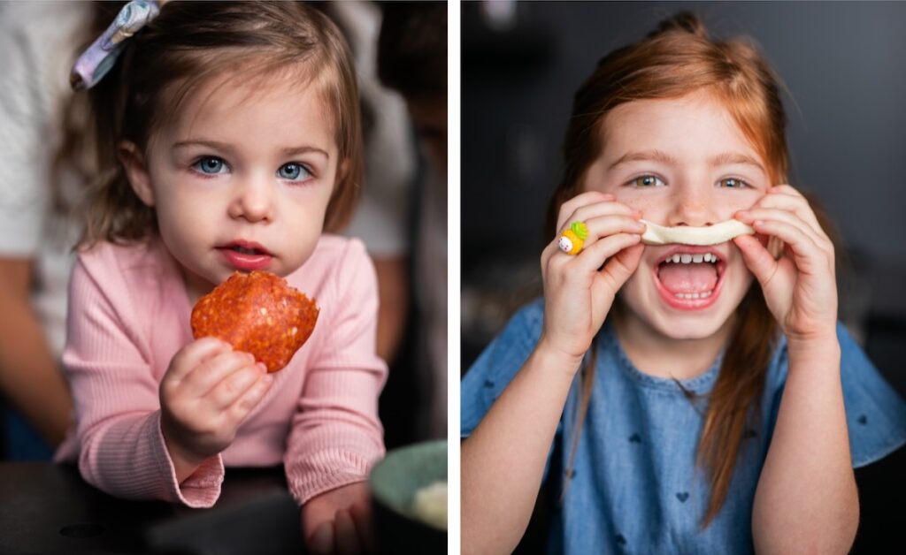 Side by side photos of two young girls, the one on the left holds a piece of pepperoni and the other holds a piece of cheese over her lip. 