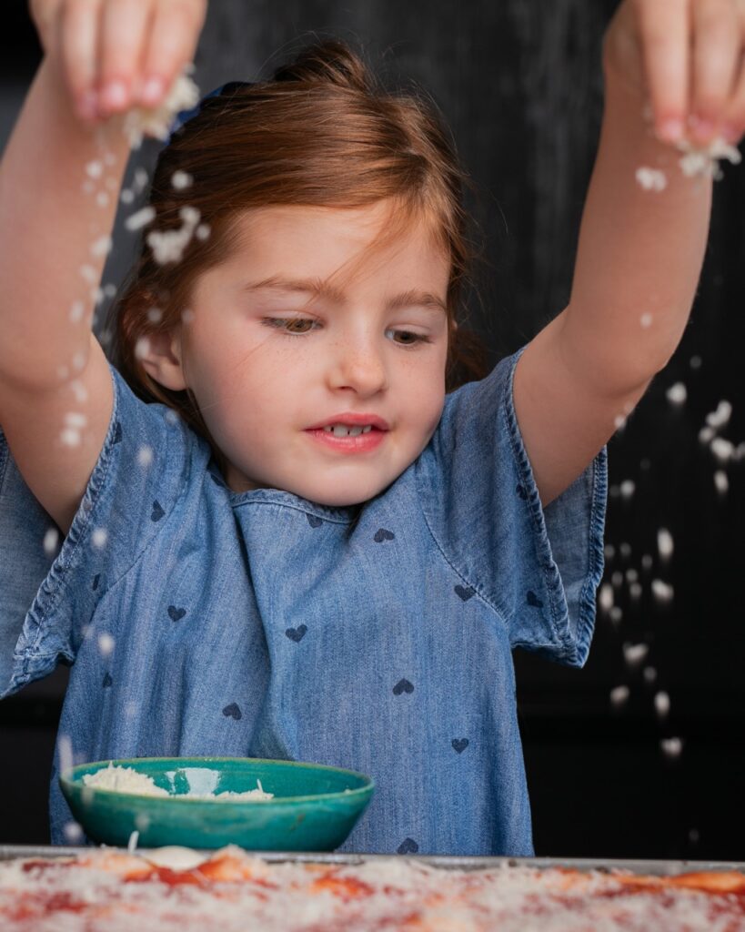 A little girl in a blue shirt sprinkles cheese onto a pizza crust.