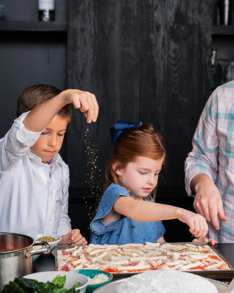 A boy and girl sibling pair sprinkle cheese on to a rectangular pizza dough.