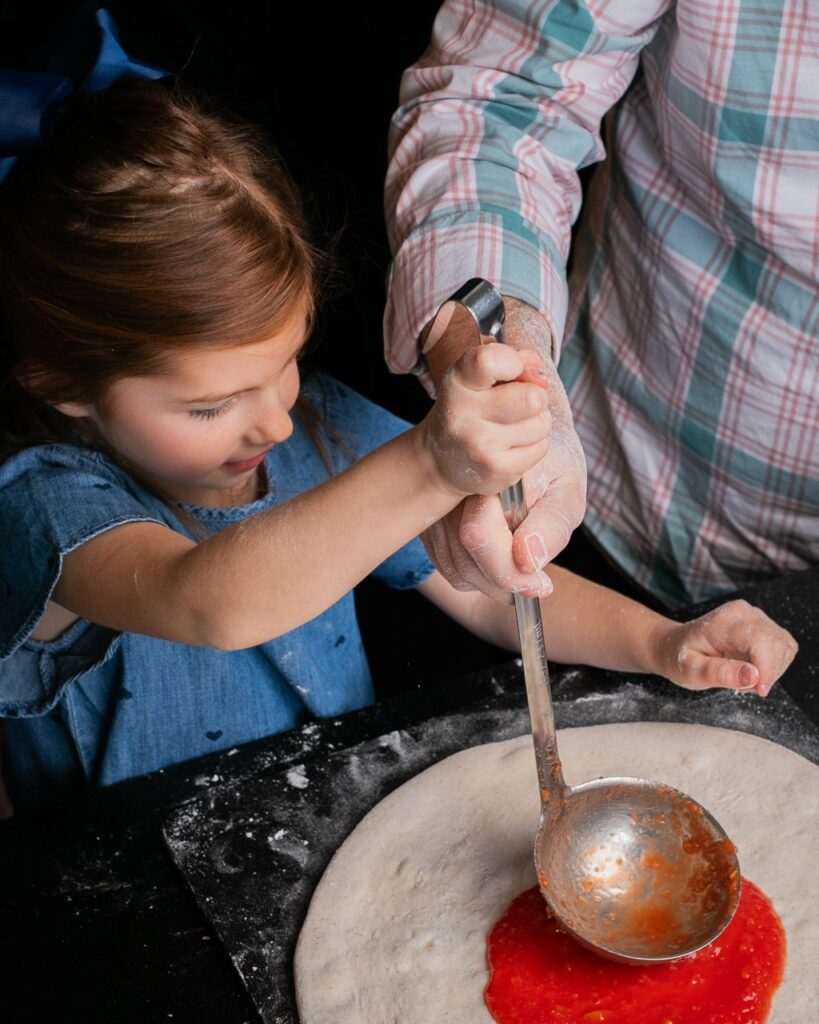 A young girl uses a ladle with her dad's assistance to spread out a red sauce on a pizza crust.