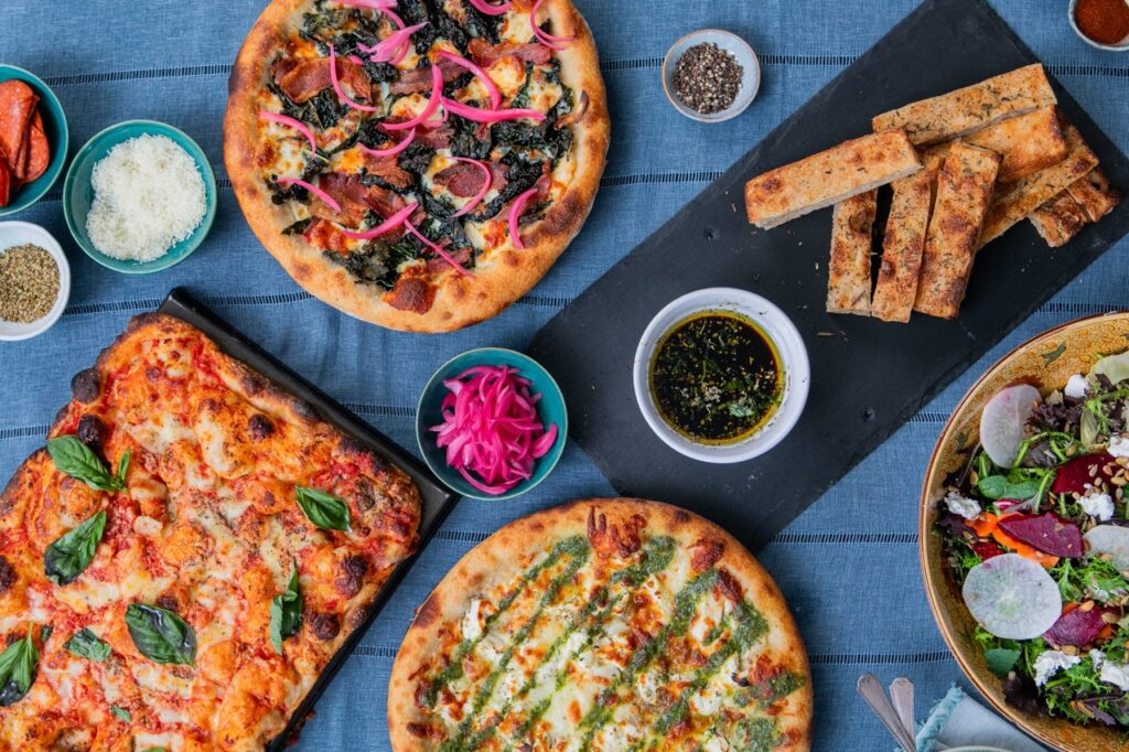 Two circle pizza, one rectangular pizza, breadsticks, and a bowl of salad sit amongst a table of sauces.