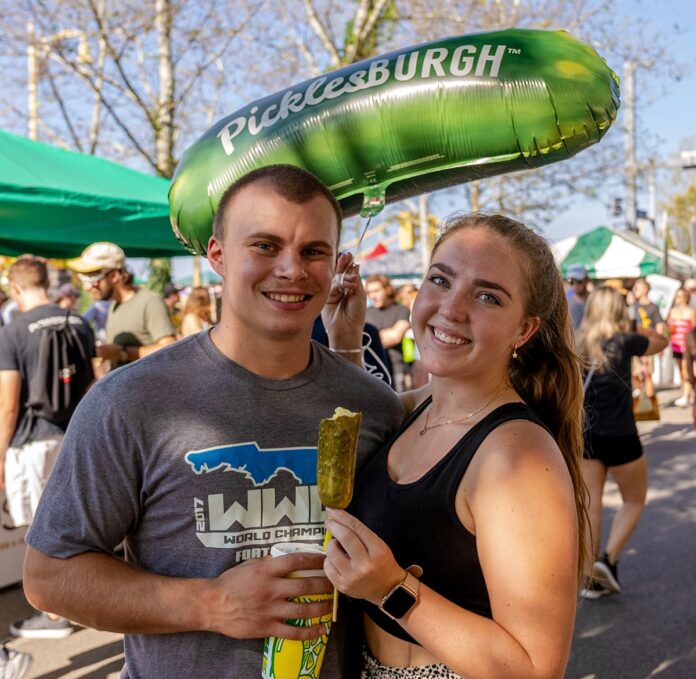Picklesburgh TABLE Magazine A man and woman stand with a pickle and lemonade in hand as a green Picklesburgh balloon floats behind them.