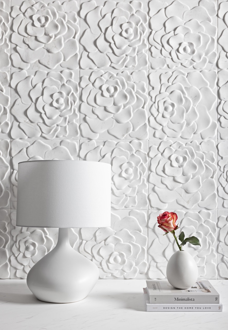 A white lamp and vase sit on a white table in front of a white, flower textured wall. 