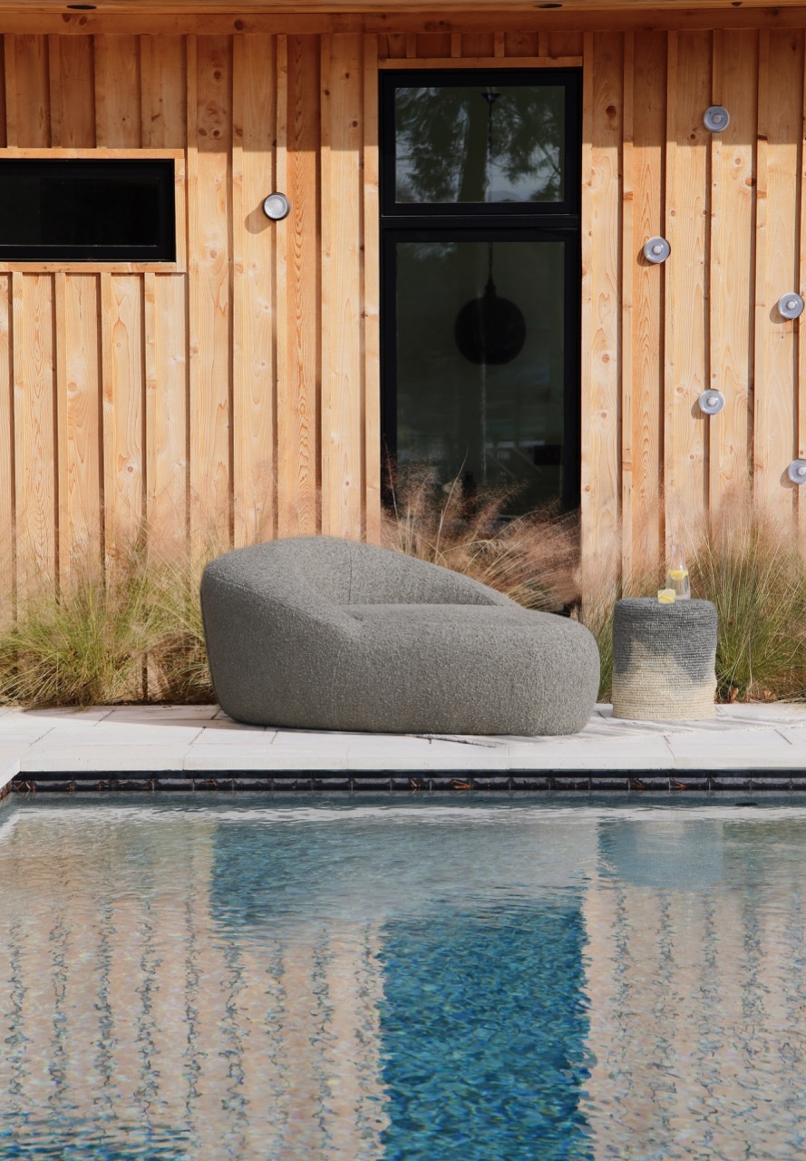 A grey lounge chair sits behind a pool and in front of a wooden shed. 