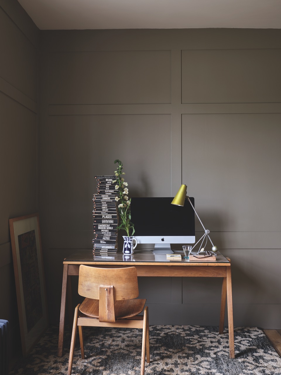 A wood desk sits against a grey back wall with a computer, lamp, and small tree on the desk.