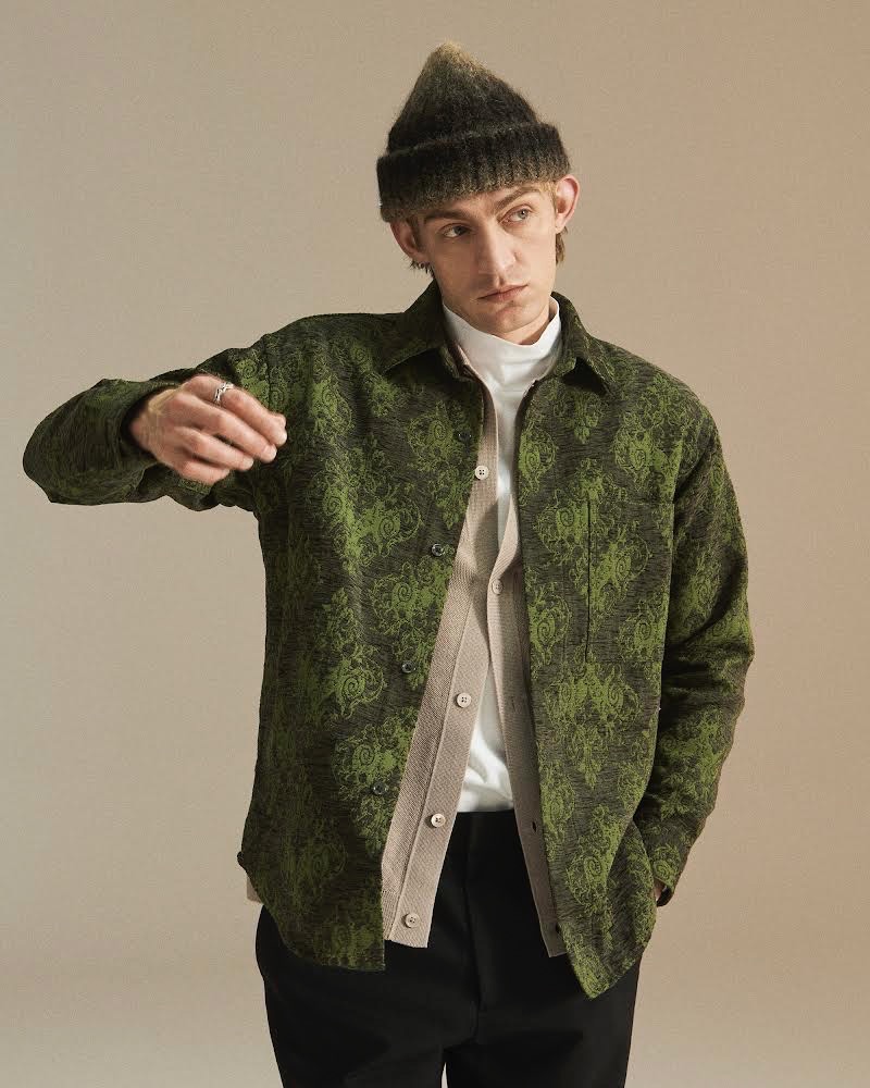 A man in a green jacket and beanie hat stands against a neutral backdrop. 