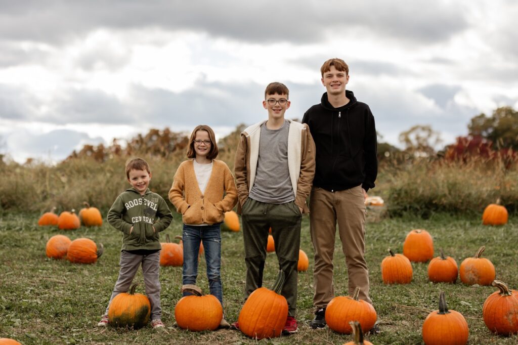 Four kids stand behind pumpkins in a pumpkin patch.