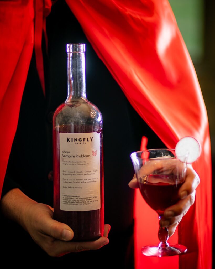 A person in black and a red cape holds a bottle and glass of Kingfly's Vampire cocktail.
