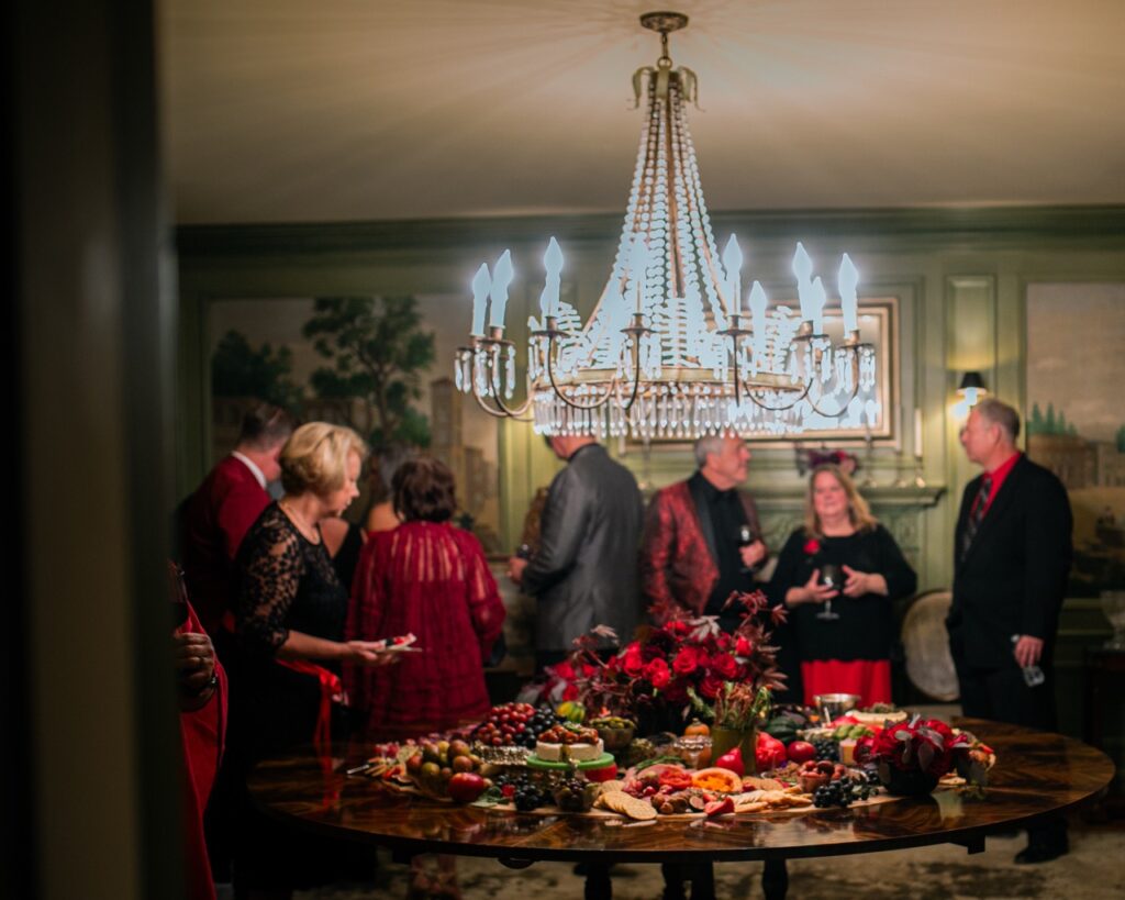 A group of guests pick up food items from a table as a chandelier hangs above their heads.