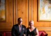 Hosting a Halloween Party Inspired by a Local Novelist’s Book A couple dressed in all black with red accents sits on a red couch in a dramatically lit wooden room.