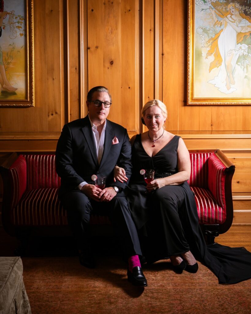 A couple dressed in all black with red accents sits on a red couch in a dramatically lit wooden room.