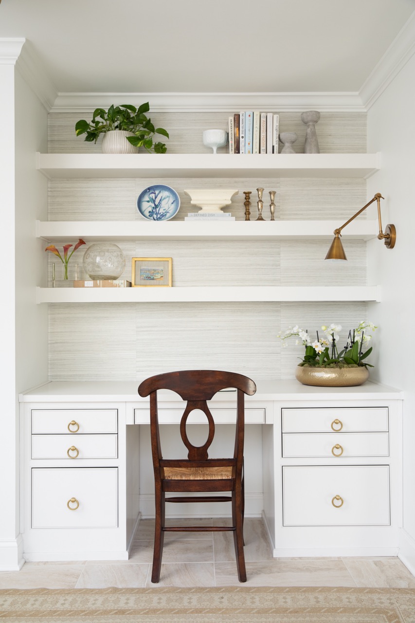 A white desk has a brown wooden chair sitting at it and white shelves above it with books and other decor. 